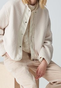 Kremowa, puszysta bomberka z futra z gładkim, jasnym zamkiem błyskawicznym z przodu. Zestawiona z teksturowanymi beżowymi spodniami z korduroju.