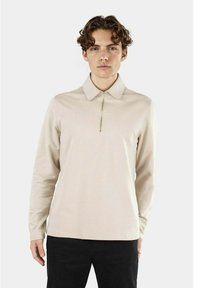 Polo a maniche lunghe beige con colletto con zip, tessuto liscio e design aderente. Indossato con pantaloni neri. Sfondo neutro.