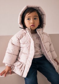 Veste matelassée rose pâle avec des motifs d'étoiles dorées et un ourlet à volants, portée par-dessus un haut à fleurs, associée à un jean en denim foncé. Capuche relevée.