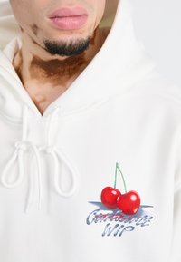 Hvit hettegenser med snøring, med et grafisk trykk av to røde kirsebær og "Carhartt WIP" i stilig skrift under.