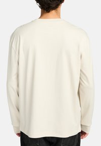 Camicia a maniche lunghe in beige chiaro, realizzata in morbido cotone. Presenta un colletto tondo e una vestibilità ampia, senza motivi visibili né dettagli in metallo.