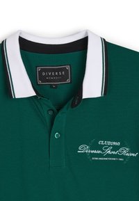 Zielona koszulka polo z białą i czarną paskowaną kołnierzykiem, dwoma guzikami, teksturowanym materiałem oraz haftowanym logo z napisem "Diverse Sport Resort."