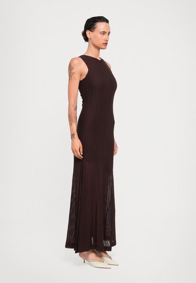 Brown ribbed long dress with a fitted bodice and floor-length hem. Made from a stretchy fabric. Paired with cream slip-on shoes.