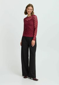 Top lungo a maniche raglan in pizzo bordeaux con scollatura rotonda abbinato a pantaloni neri a gamba larga e scarpe con tacco, creando un effetto di texture stratificata.