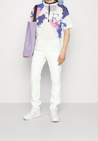 Grafická trička s modrým a růžovým skvrnitým designem, v kombinaci s bílými džíny a bílými teniskami. Fialová kabelka je přehodněná přes rameno.