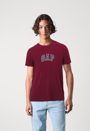 Bordeauxfarbene BaumwollT-Shirt mit einem weißen gestickten "GAP"-Logo. Rundhalsausschnitt mit kurzen Ärmeln, kombiniert mit hellblauen Jeans.
