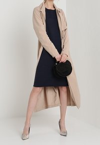 Beige trench coat over a navy dress, paired with a round black bag and patterned silver heels. The outfit features a relaxed fit and layered look.