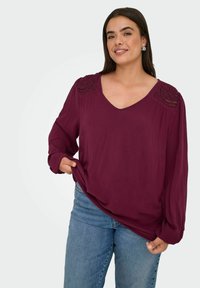 Bordeauxrote Bluse mit V-Ausschnitt, langen Ärmeln und strukturierten Spitzenakzenten an den Schultern, getragen zu hellblauen Jeans. Weicher, fließender Stoff.