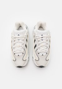 Weiße Sportschuhe mit einem glatten Lederoberteil, schwarzen Details, runder Zehenpartie und traditionellem Schnürsystem. Strukturierten Akzenten an den Seiten.