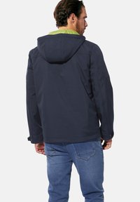 Navy jacket with a hood, green inner lining, smooth texture, and adjustable cuffs. Worn with blue jeans, viewed from the back.