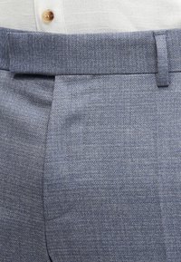 Κοντινό πλάνο στην περιοχή της μέσης με ανοιχτό μπλε υφασμάτινο παντελόνι φόρμας, που φοριέται με μπεζ πουκάμισο με κουμπιά, το οποίο είναι μπουκαρισμένο μέσα.