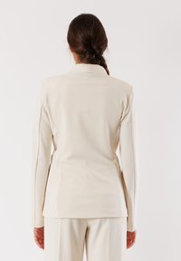 Blazer crema su misura con maniche lunghe e colletto a rotondo. Tessuto liscio con una silhouette aderente. Vista da dietro.