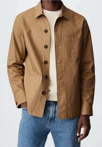 Veste marron boutonnée avec un col structuré, deux poches avant et un tissu texturé. Portée par-dessus un pull léger et un jean bleu.