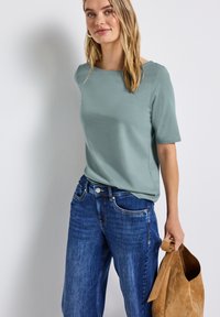 Muntgroene top met korte mouwen gecombineerd met blauwe losse jeans. Het model houdt een tan suède tas vast en toont een casual, ontspannen look.