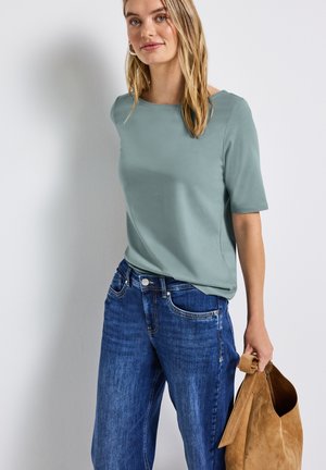 Top corto maniche corte verde menta abbinato a jeans blu a vestibilità ampia. La modella tiene in mano una borsa in suede color marrone chiaro, mostrando un look casual e rilassato.