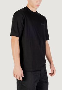 Camiseta de algodón negra de manga corta, con cuello redondo y una etiqueta texturizada en el pecho. Se lleva con pantalones negros de corte relajado.