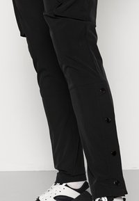 Svarta byxor med tryckknappar som går längs sidan av underbenet, bärs med vita strumpor och svart-vita sneakers på ett grått golv.