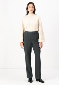 Světle béžový žebrovaný svetr s vysokým límcem a balónovými rukávy, zkombinovaný s tmavě šedými elegantními kalhotami. Model má na sobě černé kotníkové boty.