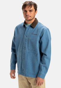 Blue denim jacket with a brown corduroy collar, button front, chest pocket, and two large front pockets. Soft texture and casual design.