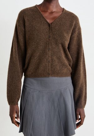 Kvinde iført en brun, blød V-hals cardigan med knapper og en højtaljet grå plisseret nederdel, stående mod en ensfarvet hvid baggrund.