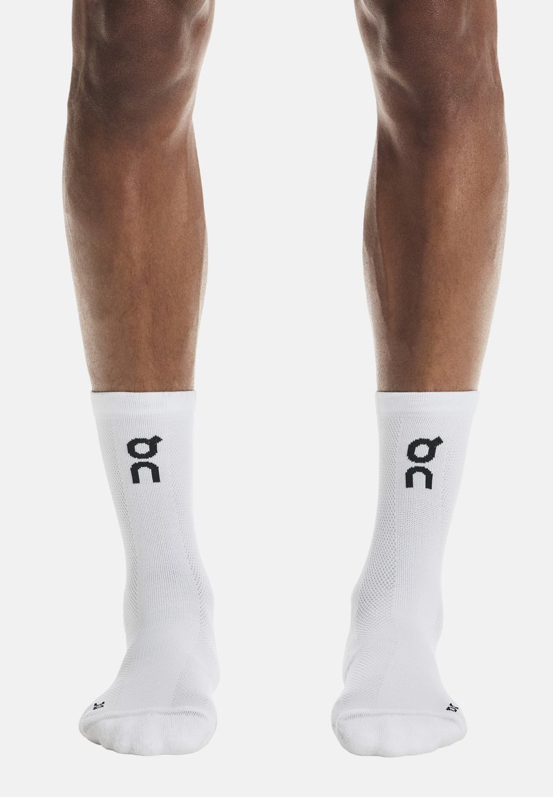 Jambes portant des chaussettes blanches mi-mollet avec un logo noir "on" de chaque côté, debout sur un fond blanc uni.