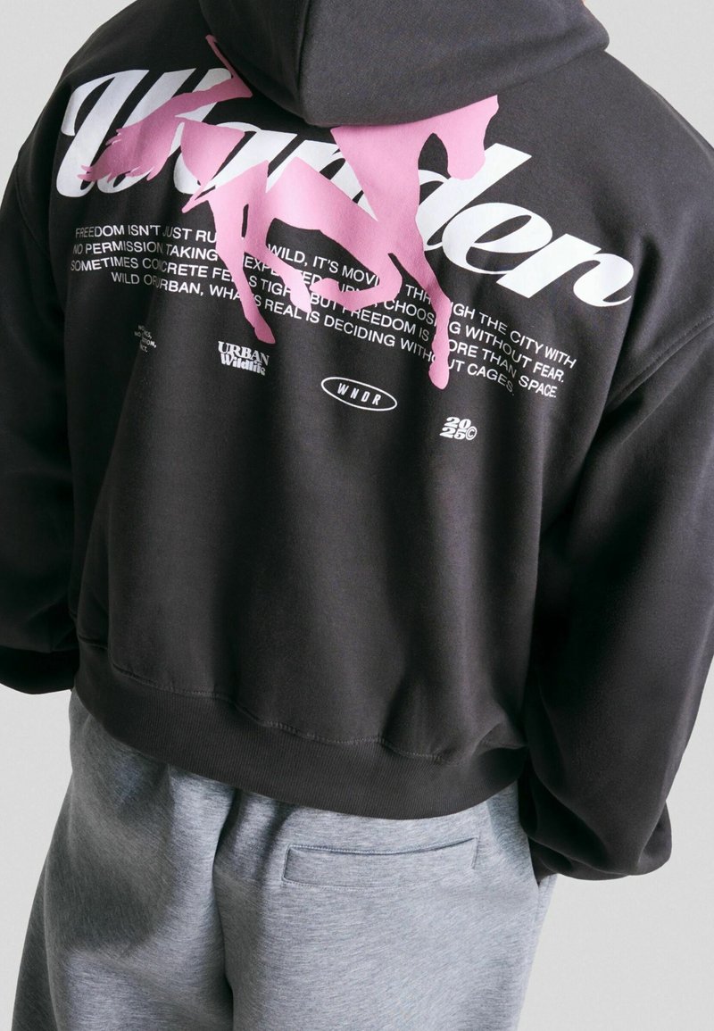 Personne portant un sweat à capuche noir avec un grand graphisme de cheval rose et un texte blanc dans le dos, associé à un pantalon gris clair.