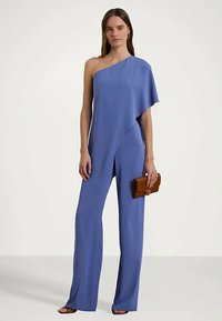 Γυναίκα που στέκεται φορώντας ένα μπλε ολόσωμο συνολάκι με έναν ώμο, κρατώντας μια καφέ τσάντα χειρός, και φοράει σκουλαρίκια κρίκους και σανδάλια.