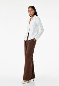 Camisa blanca de botones con cuello, de mangas largas, combinada con pantalones de pierna ancha marrones y tacones blancos de punta. Posee una postura frente a un fondo neutro.