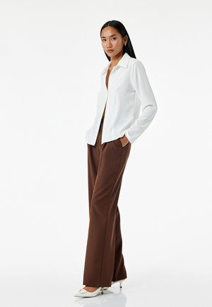 Witte blouse met kraag en lange mouwen, gecombineerd met bruine broek met wijde pijpen en witte spitse hakken, staande tegen een neutrale achtergrond.