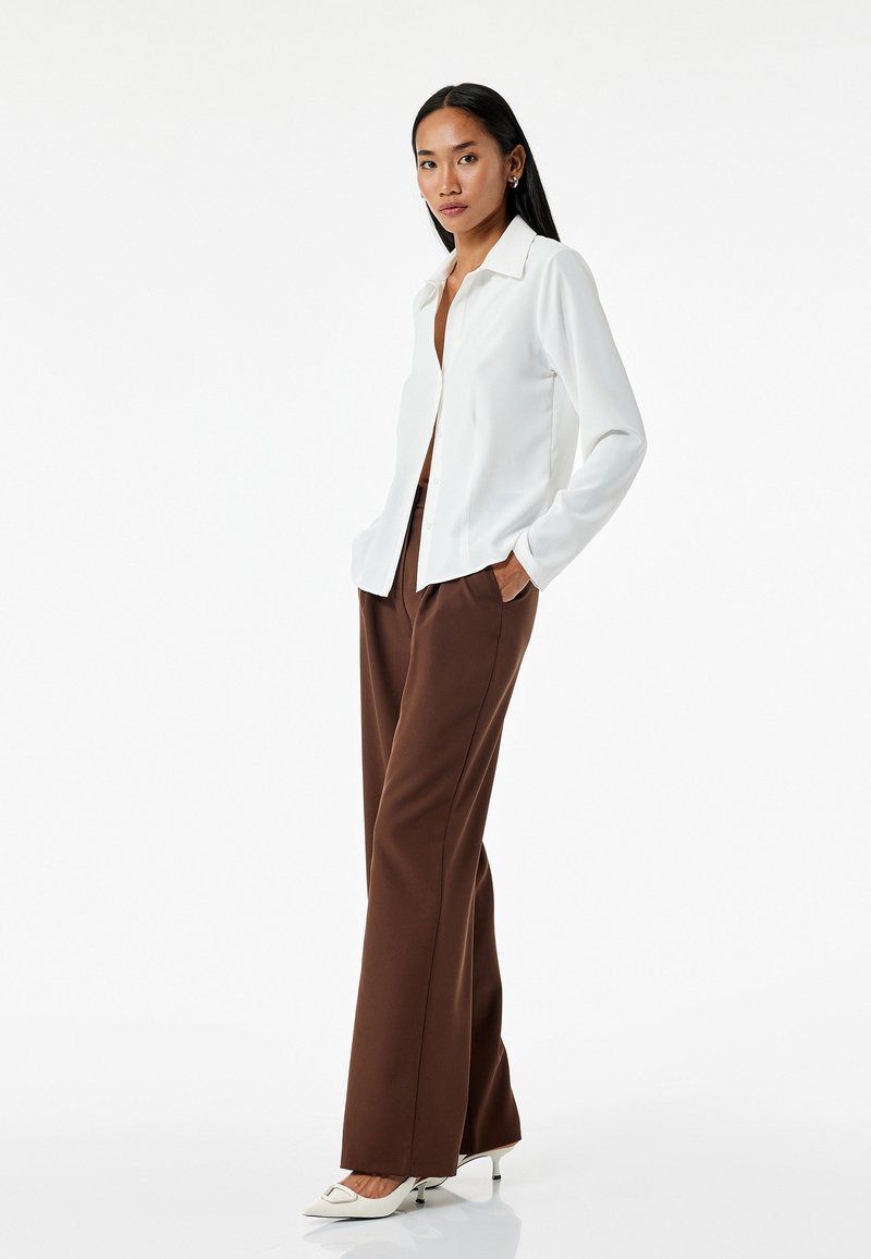 Camisa blanca de botones con cuello, de mangas largas, combinada con pantalones de pierna ancha marrones y tacones blancos de punta. Posee una postura frente a un fondo neutro.