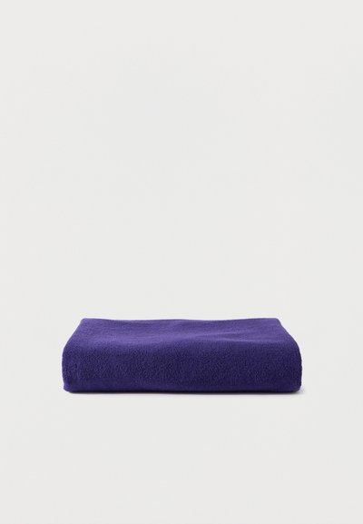 Διπλωμένη πετσέτα από πενιέ ύφασμα σκούρου μωβ χρώματος, τοποθετημένη σε φωτεινή γκρι επιφάνεια και φόντο.