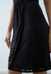 Black dress featuring a smooth fabric at the top and a lace panel with floral patterns at the bottom, with a slightly gathered waist.