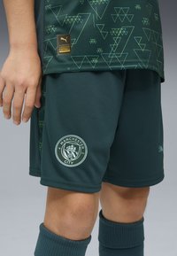 Gröna Manchester City fotbollsshorts i texturerat tyg, med en vit klubblogga och ett geometriskt mönster i ljusare grönt.