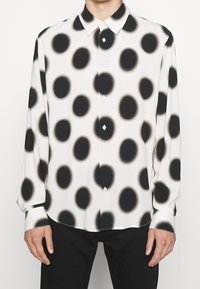 Langärmeliges Hemd aus leichtem Stoff mit weißem Hintergrund und großen schwarzen Punkten. Geknöpfter Kragen, Knopfleiste vorne und schwarze Akzente.