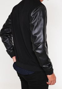 Chaqueta bomber negra con cuerpo de tela y mangas de cuero. Dobladillos y puños elásticos. Se lleva sobre una camisa azul marino, que presenta un cuello redondeado.