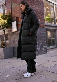 Giubbotto a vento nero con un design lungo e sovradimensionato, dotato di colletto alto, chiusura con zip e grande motivo a quadri rettangolari.