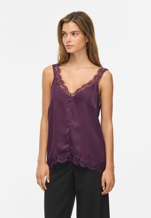 Femme portant un top en satin violet sans manches avec une bordure en dentelle au niveau de l'encolure et de l'ourlet, assorti à un pantalon noir, debout devant un fond blanc.