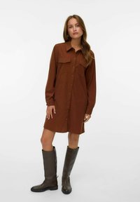 Robe chemise à manches longues et à boutons en tissu couleur rouille, dotée de deux poches poitrine, associée à des bottes marron jusqu'aux genoux.
