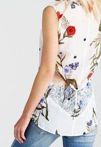 Blouse sans manches blanche avec imprimé floral rouge, bleu et vert ; présente des détails en dentelle à l'arrière. Tissu semi-transparent et léger.