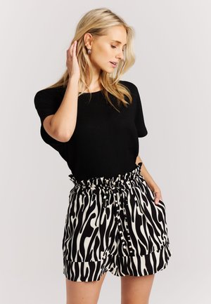 Blonde woman wearing black short-sleeve top and high-waisted black and white zebra print shorts with pockets, posing with hand near hair.