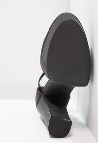 Chaussure noire à talon posée sur le côté contre une surface blanche, montrant une semelle lisse et un talon arrondi.