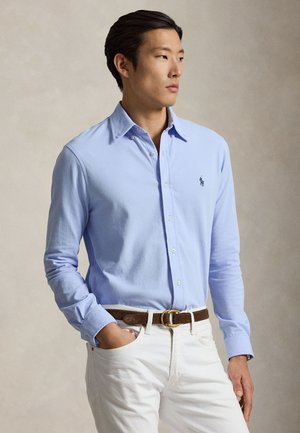 Ung mand med mørkt hår iført en lyseblå skjorte med knapper, hvide bukser og brunt bælte, ser let mod højre.