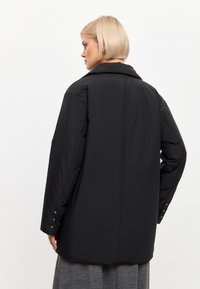 Chaqueta de gran tamaño negra con un cuello estructurado, textura acolchada y detalles de botones en las mangas. Tejido suave, diseño minimalista.