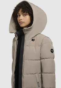 Chaqueta acolchada beige con una gran capucha, cierre de cremallera y una franja de acento negra. Presenta un logo redondo en la manga izquierda y bolsillos frontales.