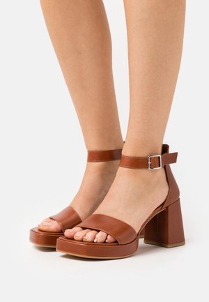 Brune læderplatformsandaler med en bred tårem, ankelrem med en sølvspænde og en kraftig blokhæl. Teksturop og glatte overflader.