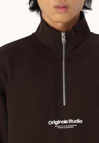 Sudadera marrón con cremallera de cuarto con un cuello alto y cremallera plateada. Presenta el logo "Originals Studio" en blanco en la parte delantera. Textura suave.