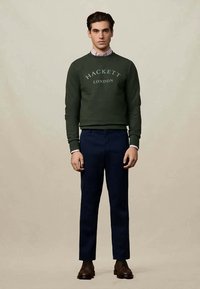 Homem a standing wearing a sweatshirt verde da Hackett London, camisa riscada, calças azul-marinho e sapatos de couro castanho sobre um fundo bege liso.