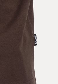 Brązowa bawełniana koszulka z gładką teksturą i małą, czarną metką z logo wyszytą na boku, charakteryzująca się minimalistycznym wzornictwem.