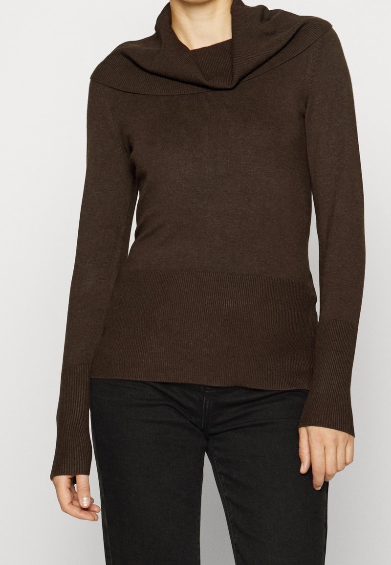 Femme portant un pull en maille marron à manches longues avec un large col large rabattable et un pantalon noir, debout devant un arrière-plan uni.