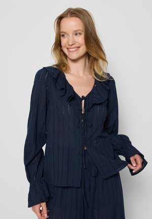 Navyblauwe blouse met rucheskraag, lange mouwen en striksluiting. Heeft een opengewerkt patroon en een gestructureerde stof. Losse pasvorm.